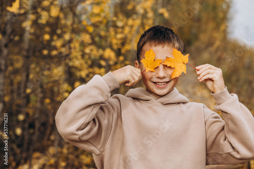 Teenager boy hiding his eyes behind maple leaves. Child holding yellow autumn leaves in his hands. Teen having fun on walking in autumn park. Selective focus