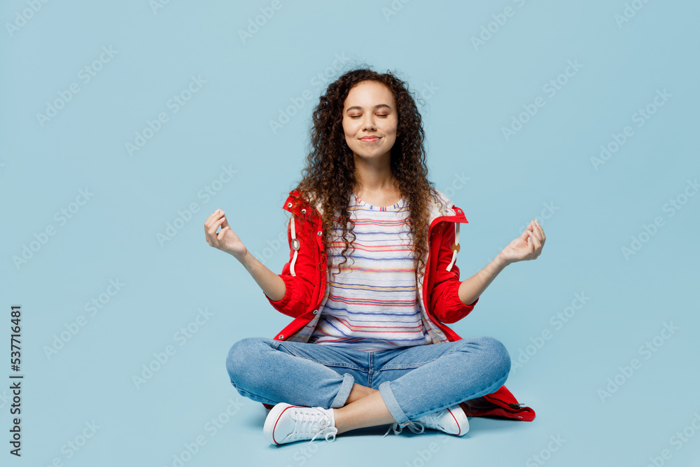 Full body young woman of African American ethnicity wear red jacket sit hold spreading hands in yoga om aum gesture relax meditate try to calm down isolated on plain pastel light blue cyan background. Full body young woman of African American ethnicity wear red jacket sit hold spreading hands in yoga om aum gesture relax meditate try to calm down isolated on plain pastel light blue cyan background.