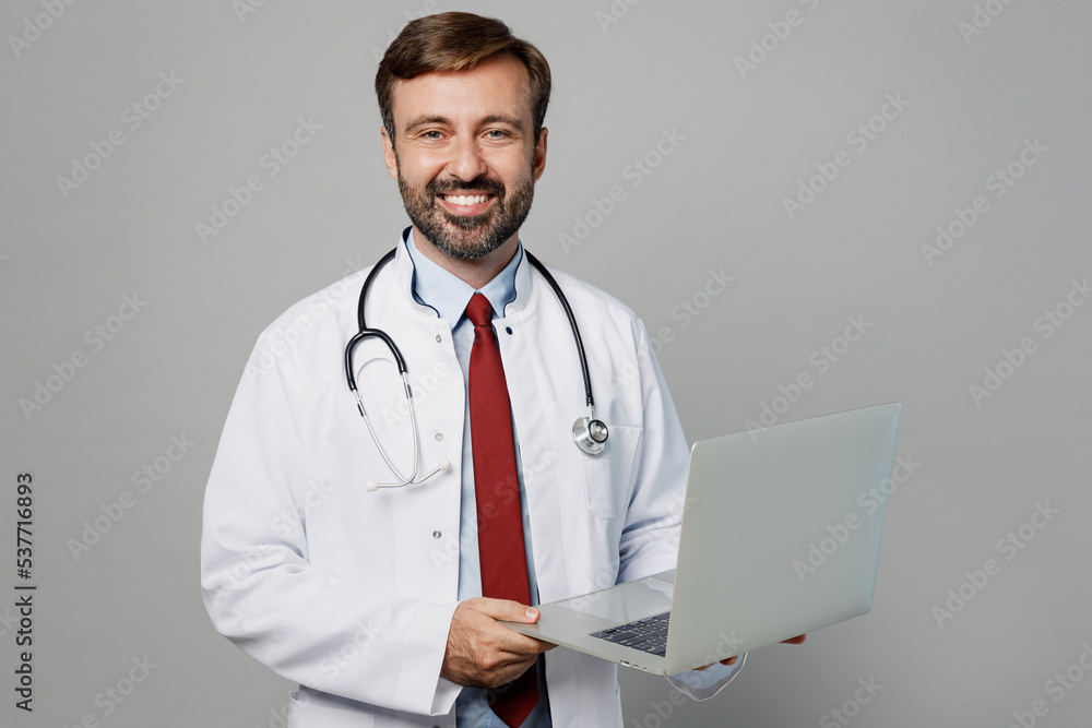 Male doctor smiling IT man wears white medical gown suit stethoscope work in hospital hold use laptop pc computer isolated on plain grey color background studio portrait. Healthcare medicine concept. Male doctor smiling IT man wears white medical gown suit stethoscope work in hospital hold use laptop pc computer isolated on plain grey color background studio portrait. Healthcare medicine concept.