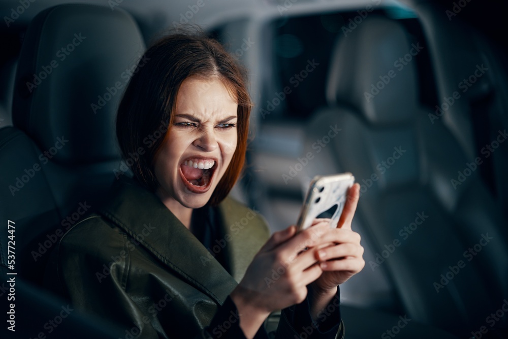 a close horizontal portrait of a stylish, luxurious woman in a leather coat sitting in a black car at night in the passenger seat, emotionally screaming while holding her smartphone