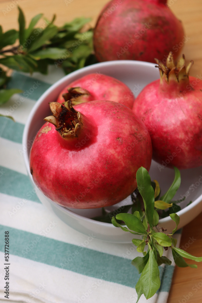 Bright red Pomogranate fruits in a white bowl on wooden table with green leaves. Punica granatum fruit Bright red Pomogranate fruits in a white bowl on wooden table with green leaves. Punica granatum fruit