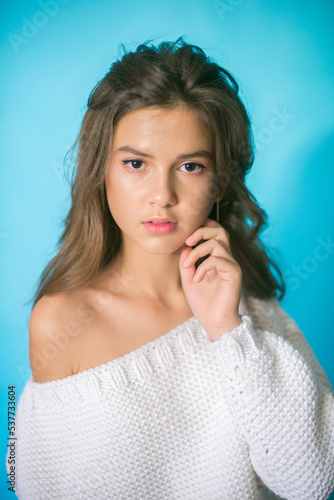 
A cute teenage girl with long dark hair in trendy jeans and a white knitted sweater on a blue background in the studio. Fashion for teenagers