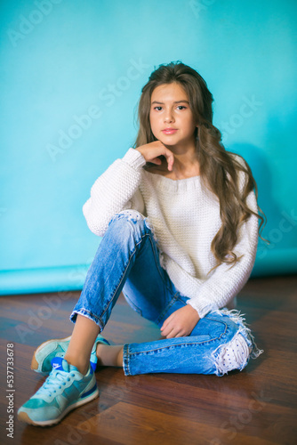 
A cute teenage girl with long dark hair in trendy jeans and a white knitted sweater on a blue background in the studio. Fashion for teenagers