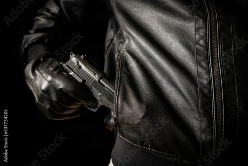 concept of the crime of banditry. A dangerous shooter and a black pistol on a dark background. The hired killer is preparing to shoot. Pulls a firearm out of his jacket pocket