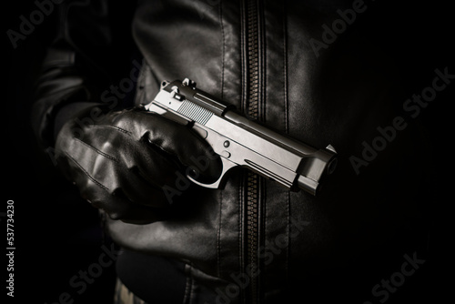 The concept of the crime of banditry. A dangerous shooter and a black pistol on a dark background. hired killer is preparing to shoot. Pulls a firearm out of his jacket pocket