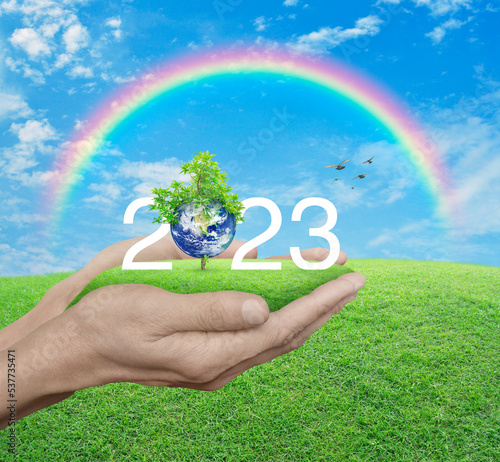 2023 white text with planet and tree on green grass in hands over blue sky and rainbow, Happy new year 2023 ecological cover, Save the earth concept, Elements of this image furnished by NASA
