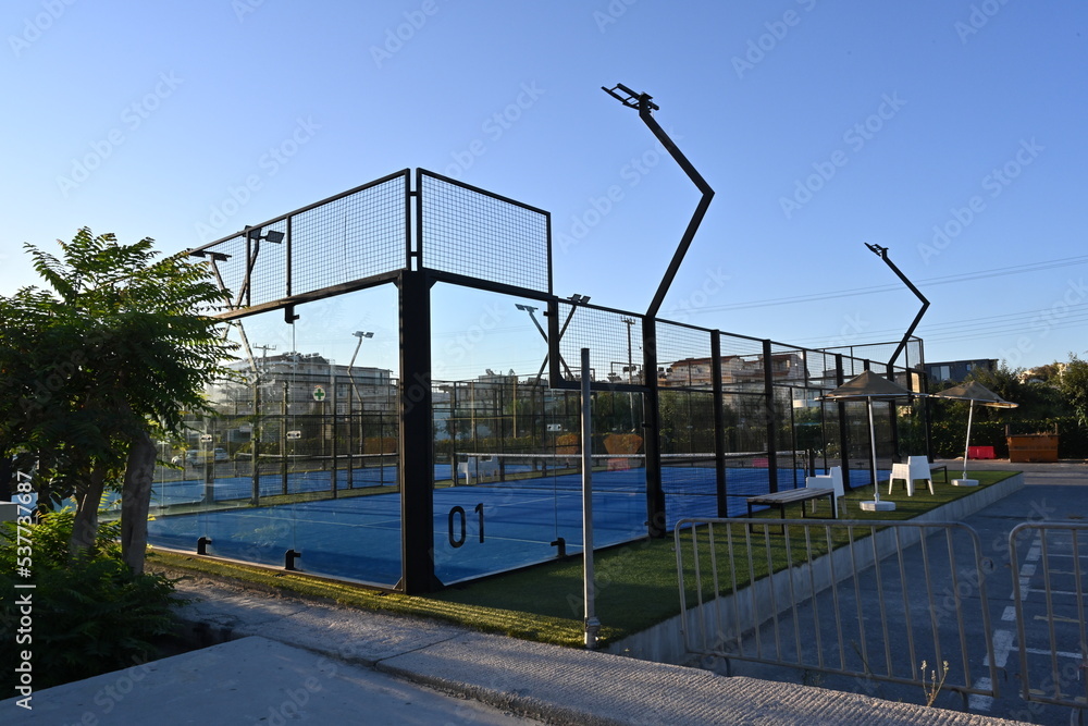 An enclosed blue court for padel with construction created by mesh and the glass back walls. Padel is a racket sport played in doubles and is similar to tennis with the same rules of scoring. An enclosed blue court for padel with construction created by mesh and the glass back walls. Padel is a racket sport played in doubles and is similar to tennis with the same rules of scoring.