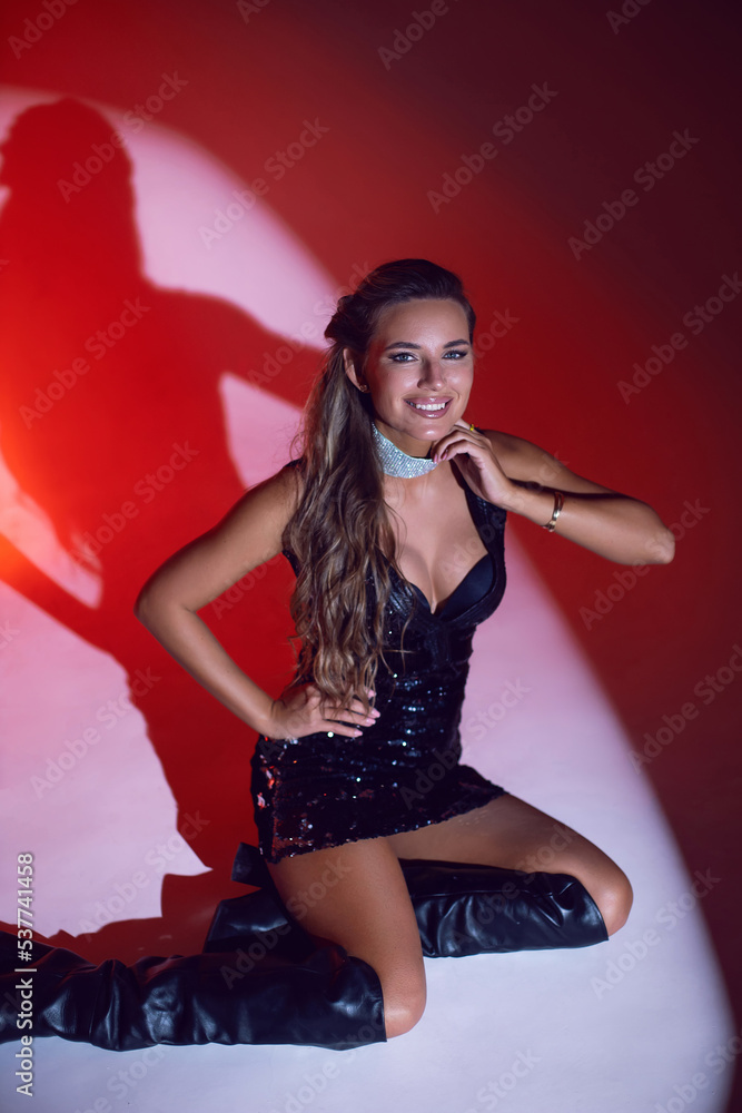 Naklejka premium portrait happy of a sexy woman in a dress and boots sit in the studio on a cyclorama on a red background .