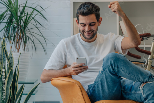фотографія young man at home euphoric celebrating looking at mobile phone