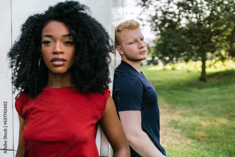 Serious multiethnic couple standing near white building in park Serious multiethnic couple standing near white building in park