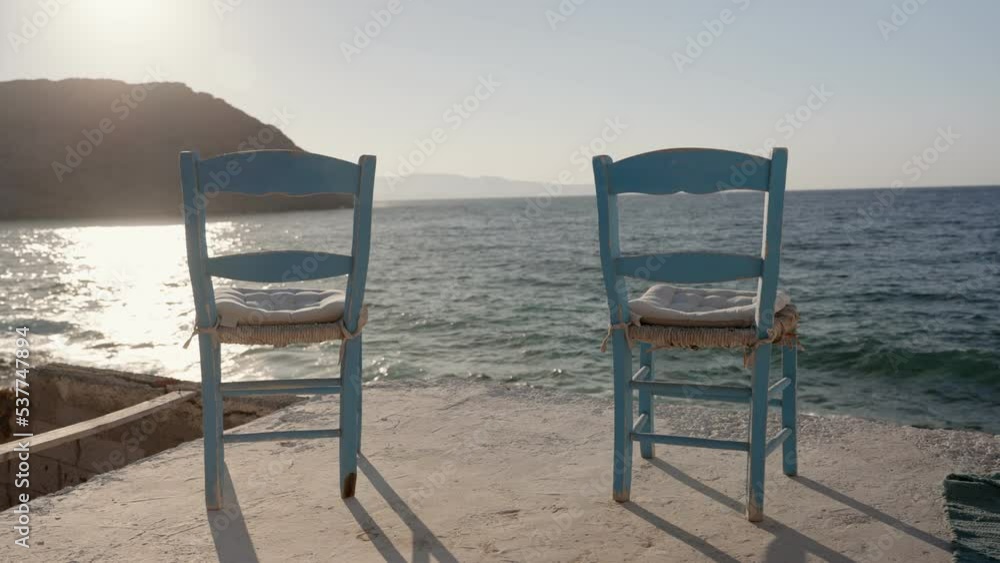 Two light blue chairs set in front of cliff and sea in afternoon sun in Greece. Set of classic taverna chairs in blue color overlooking the Mediterranean as waves crash and sun is flaring
