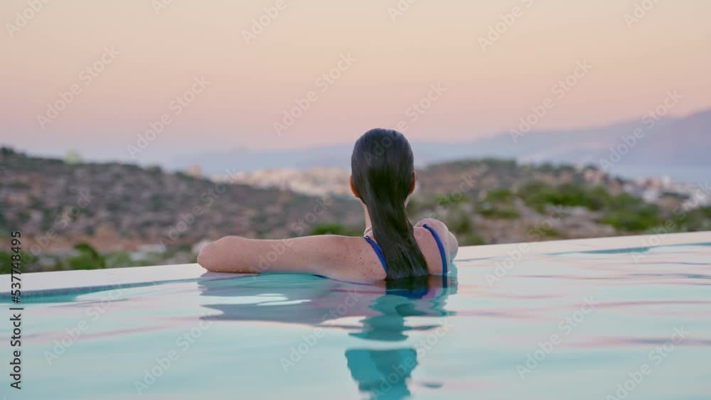 Young woman sitting in luxury infinity pool in hotel or suite at sunset ...