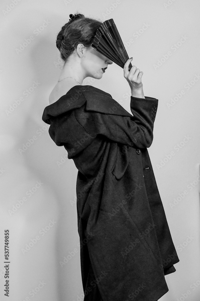 Obraz premium Vertical grayscale shot of an elegant female posing in a slip dress, trench coat, and a fan