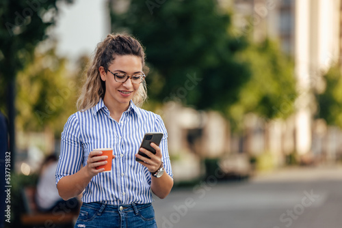 Fototapeta Naklejka Na Ścianę i Meble -  Smiling girl walking on the street, holding cup of coffee and using a phone.