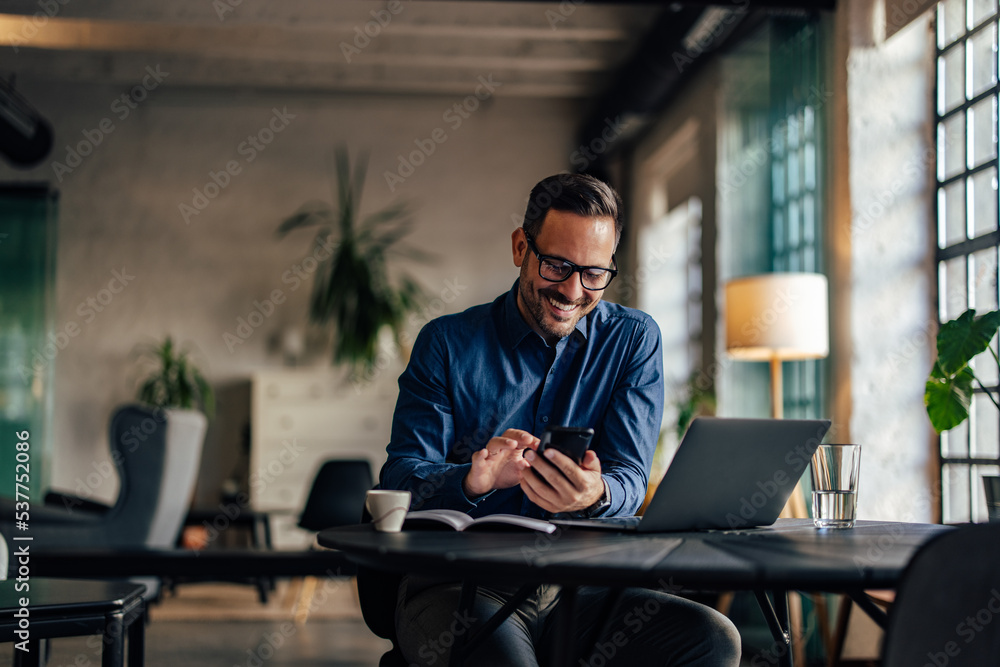 Photo of a smiling man using a mobile phone while being at the office.