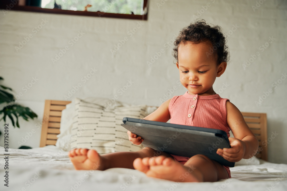 Focused biracial baby girl sitting on bed with digital tablet at home Focused biracial baby girl sitting on bed with digital tablet at home