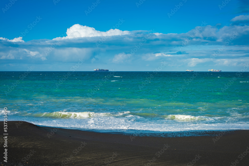 Sea waves breaking on a black sand beach. Distant cargo ship sailing at the horizon