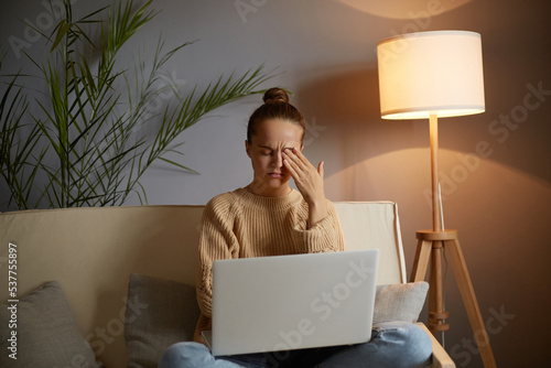 Obraz na plátně Indoor shot of tired woman wearing casual clothing sitting on sofa and working on notebook, feels pain in her eyes, rubbing eye, working for long time, felling fatigue
