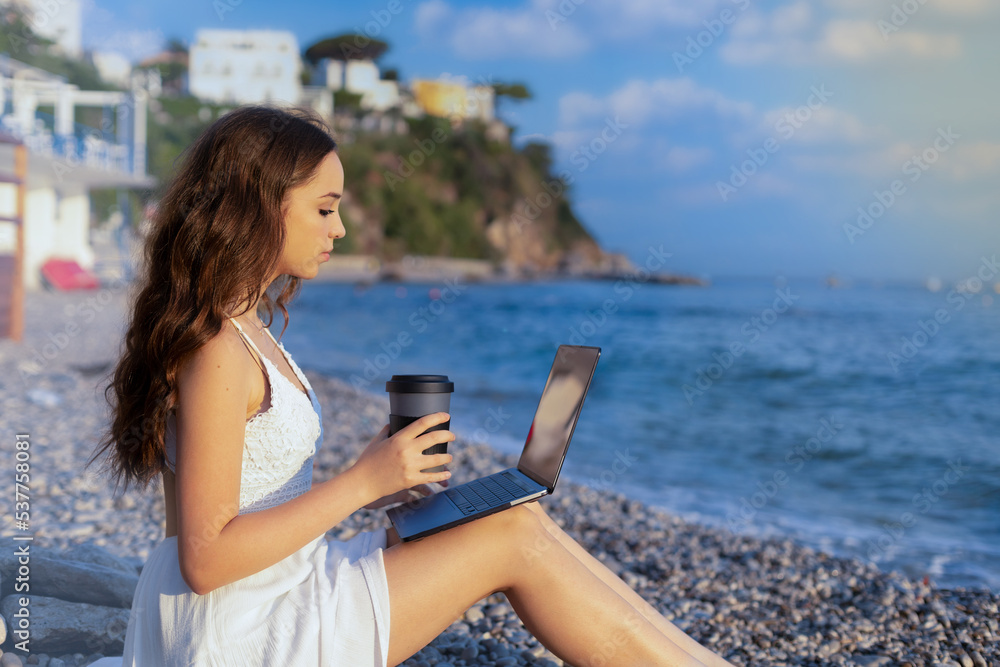 Teenage girl works on computer and talks on phone on the beach. concept of work and vacation together, digital nomads around the world