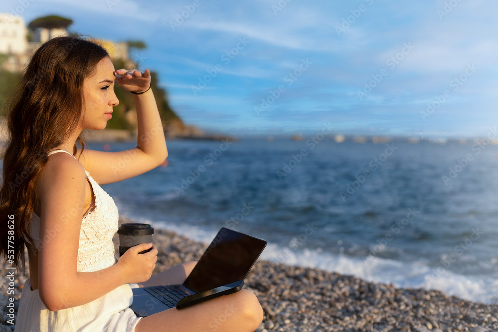 Teenage girl works on computer and talks on phone on the beach. concept of work and vacation together, digital nomads around the world