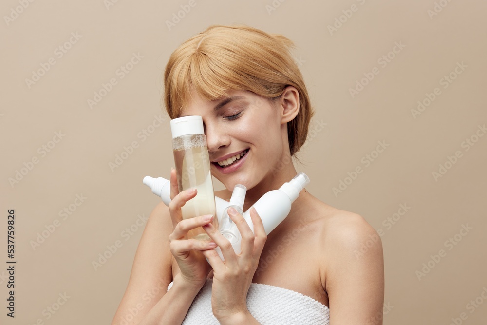 a funny, cheerful woman stands on a beige background holding various jars of care cosmetics in her hands and smiling pleasantly, taking them to her face, closes her eyes with happiness a funny, cheerful woman stands on a beige background holding various jars of care cosmetics in her hands and smiling pleasantly, taking them to her face, closes her eyes with happiness