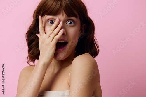 a close horizontal photograph of a surprised, admiring, attractive young woman on a pink background, with her mouth wide open in surprise, covering it with her hand