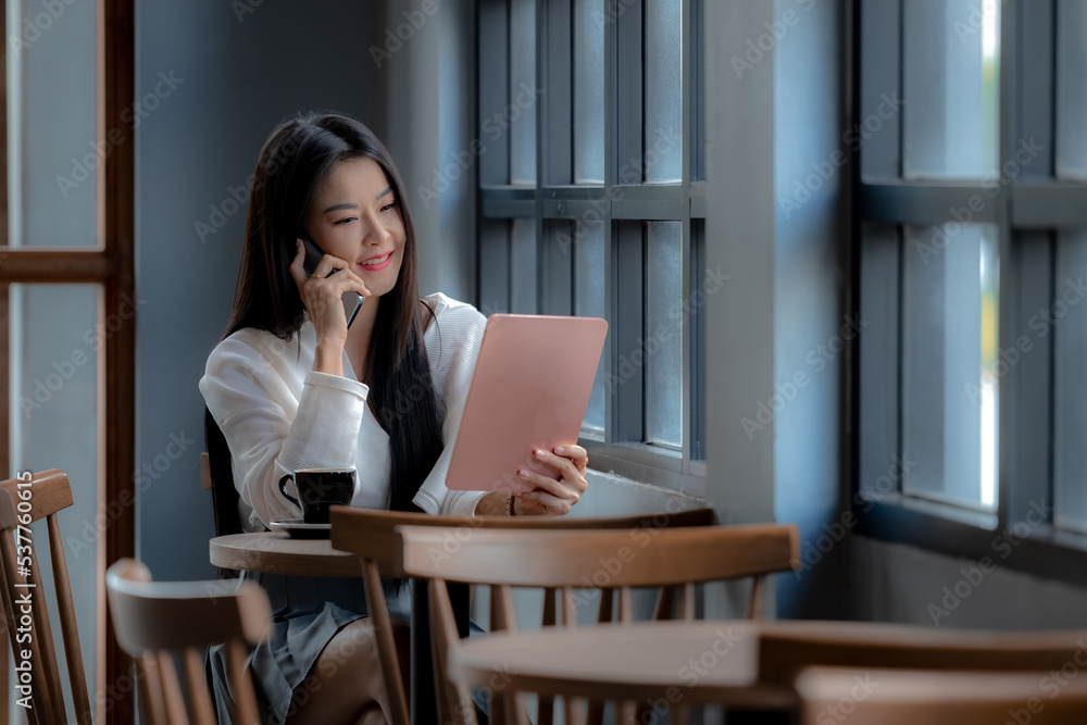Asian woman using tablet and talking conversation by mobile in cafe. Woman looking tablet and calling with mobile smartphone in cafe. Young asian woman happy face smiling and using mobile smartphone. Asian woman using tablet and talking conversation by mobile in cafe. Woman looking tablet and calling with mobile smartphone in cafe. Young asian woman happy face smiling and using mobile smartphone.