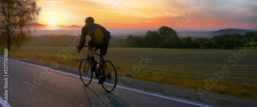  Cyclist riding his bicycle on an empty rural road at dawn, epic sunrise in the background