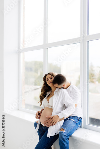 Happy man hugging his pregnant wife sitting on a big window. Happy young family sitting on the windowsill of their new home. Lovers spend time together.