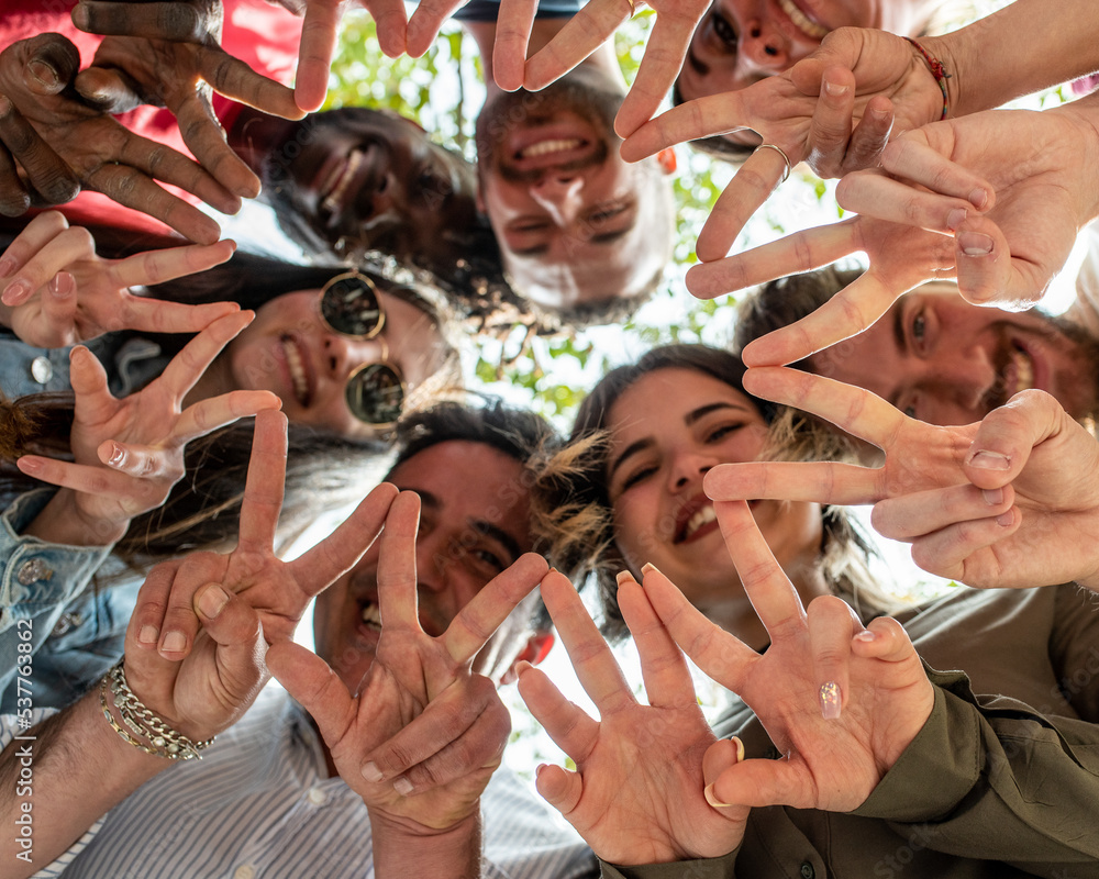 Obraz premium Group of happy friends standing in circle in park and showing peace or victory sign, view from below, seven people from a multiracial family
