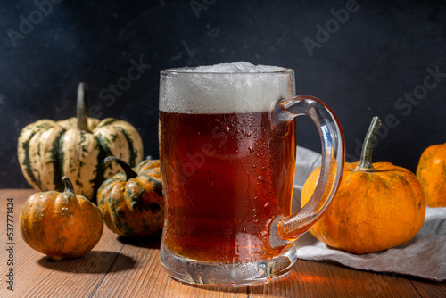 Dark frothy pumpkin ale in beer glass mug, autumn homemade alcohol drink, with small pumpkins on wooden kitchen table 