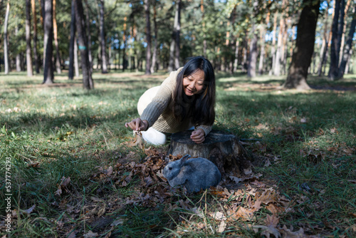 Adorable young rabbit and woman sit together outdoor in autumn forest. Asian woman wear warm sweater with pet in relax and calm.