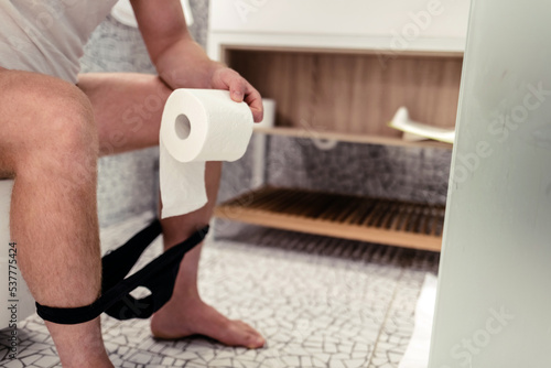 Cropped photo of Caucasian man sitting on toilet seat with roll of toilet paper. A man is sitting on a toilet in a bathroom. diarrhea. Stomach pain.