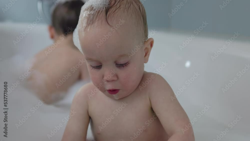 two sibling brothers take bath together having fun with soap foam playing water bubbles. baby boy curious filling bottle with water trying to eat foam. toddler boy washing hair under water tap faucet two sibling brothers take bath together having fun with soap foam playing water bubbles. baby boy curious filling bottle with water trying to eat foam. toddler boy washing hair under water tap faucet