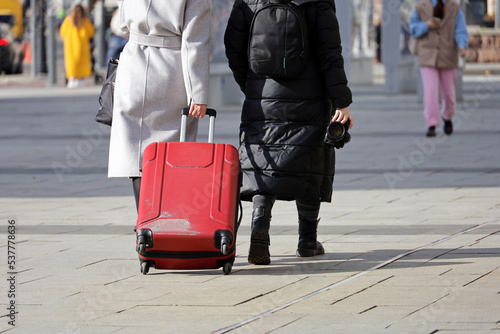 Two women wearing autumn clothes walking with red suitcase on wheels down the street. Female legs and luggage on city sidewalk, travel concept
