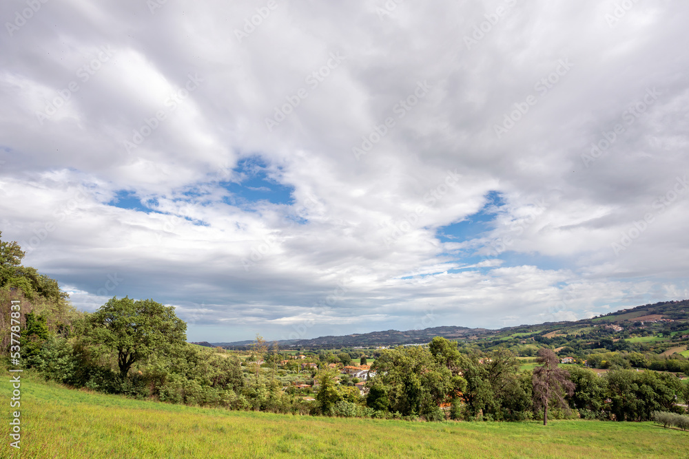 Cloudscape over the hills in the Montefeltro. The village of Case Bernardi near Pesaro and Urbino in the Marche region of Italy appears through the green trees Cloudscape over the hills in the Montefeltro. The village of Case Bernardi near Pesaro and Urbino in the Marche region of Italy appears through the green trees
