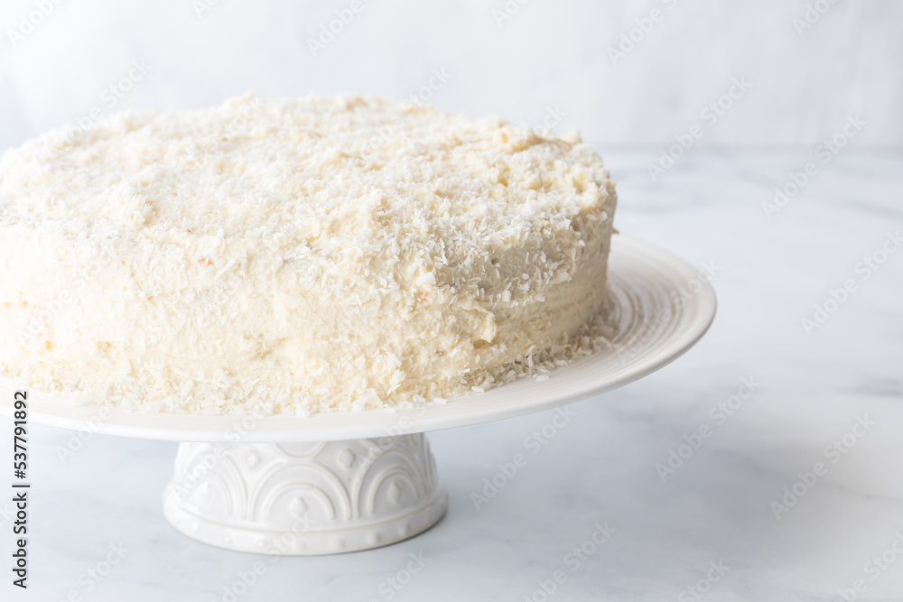 A coconut covered cake on a white pedestal stand against a white background.