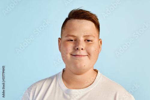 Cute smiling boy looking at camera. Teen boy in white tee expressing positive emotions isolated over blue background. Kids emotions concept