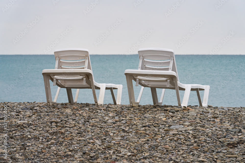 Two white plastic sun beds on a pebble beach near the sea coastline. Copy space.