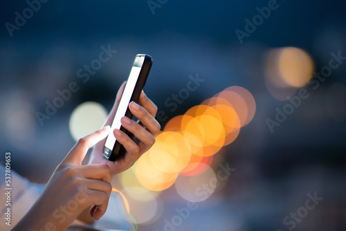 Asian woman holding smartphone blank screen on city light bokeh glowing background in night atmosphere.