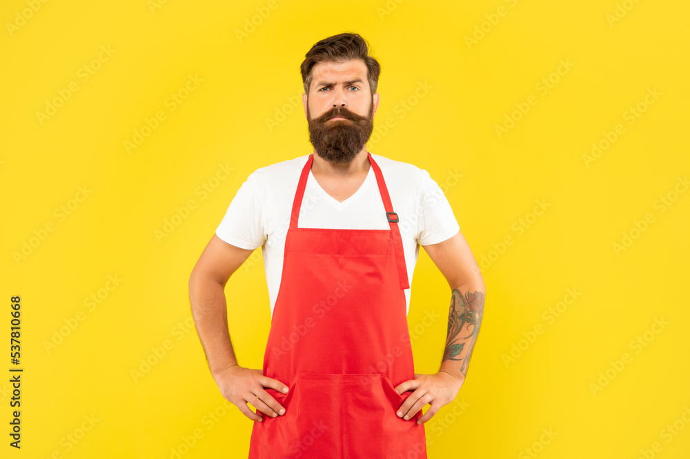 Confident serious man with arms akimbo in red apron yellow background, household Confident serious man with arms akimbo in red apron yellow background, household