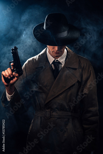 Silhouette of a male detective in a coat and hat with a gun in his hands. A dramatic noir portrait in the style of books and detective films of the 1950s.