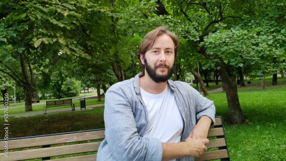 Handsome young man smiles looking at camera. Cute guy is sitting on bench in city public park on summer day. Happy person takes break from work and household chores. Portrait of man in casual clothes Handsome young man smiles looking at camera. Cute guy is sitting on bench in city public park on summer day. Happy person takes break from work and household chores. Portrait of man in casual clothes