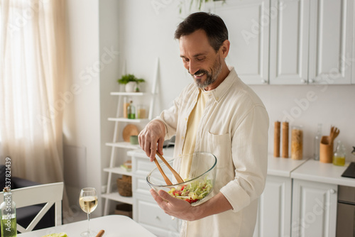 cheerful man mixing vegetable salad with wooden tongs near glass of white wine in kitchen.