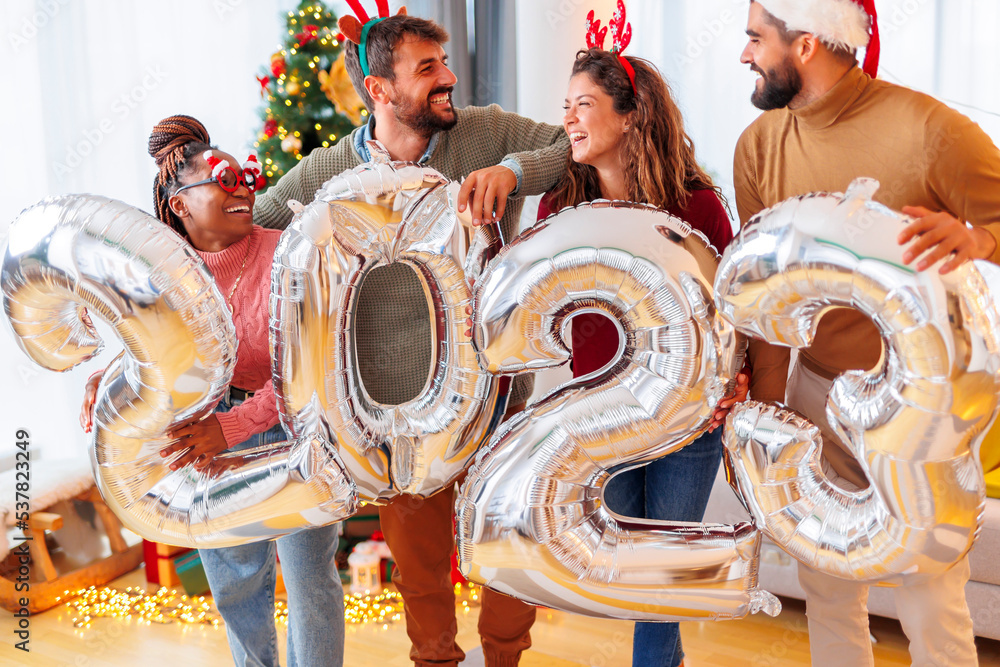 Friends holding big balloons shaped as numbers 2023 while celebrating New Year at home