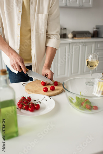 partial view of man cutting fresh cherry tomatoes near glass of white wine and blurred bottle of oil.
