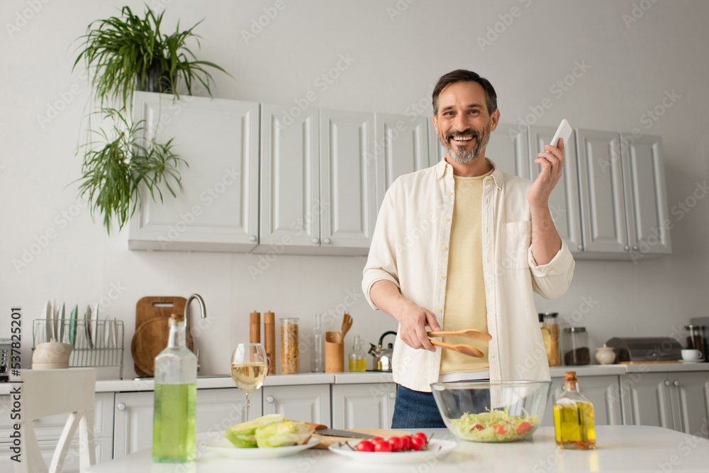 happy man with smartphone and wooden tongs looking at camera near fresh vegetables and bottles with oil. happy man with smartphone and wooden tongs looking at camera near fresh vegetables and bottles with oil.