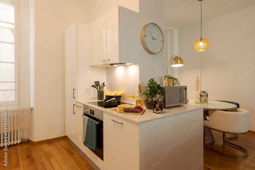 Modern white kitchen with parquet. On the fire there is the pot with the pasta and the hood light is on.