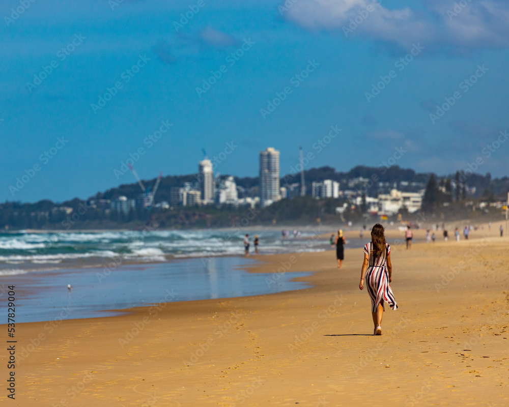 beautiful long-haired girl in long colorful dress walks along beach in gold coast on sunny day; beach with huge skyscrapers; miami beach in gold coast, australia beautiful long-haired girl in long colorful dress walks along beach in gold coast on sunny day; beach with huge skyscrapers; miami beach in gold coast, australia