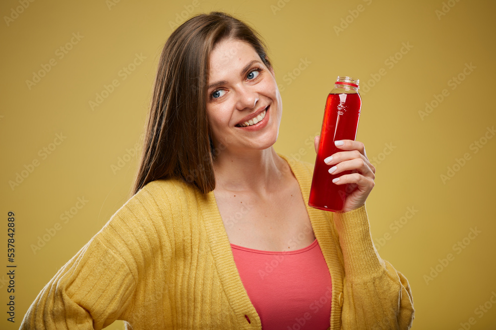 Smiling woman holding red bottle with juice drink looking away. Isolated advertising portrait on yellow. Smiling woman holding red bottle with juice drink looking away. Isolated advertising portrait on yellow.
